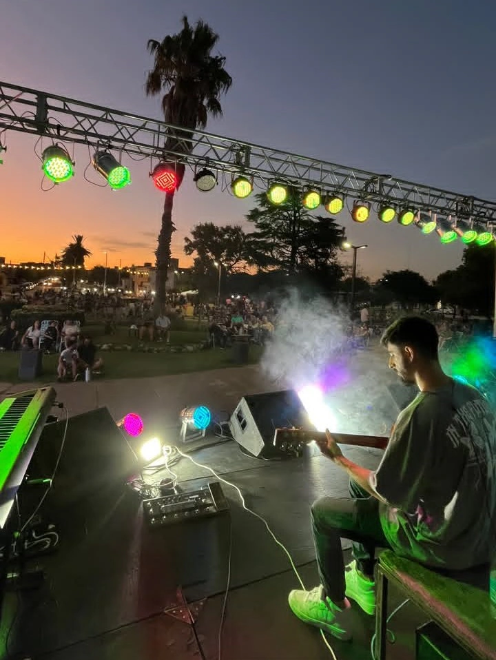 Artist playing guitar with a stage set for him, big audience in the distance with unfolding sunset